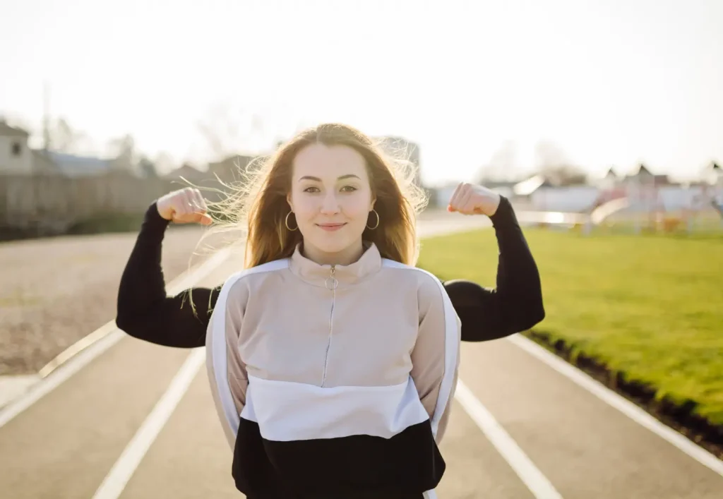 Une jeune femme au visage serein sourit, debout au milieu de la route, et contracte ses biceps, parce qu'elle a réussi l'examen du permis de conduire sans stress grâce à l'hypnose.