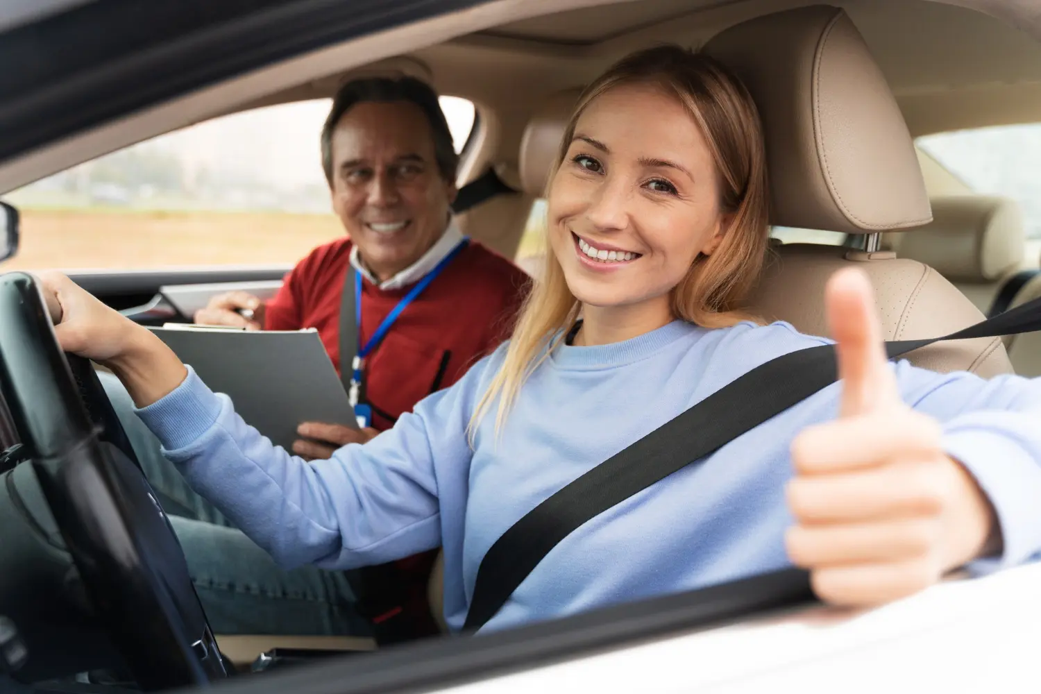 Une jeune femme au volant d'une voiture, sourire aux lèvres et pouce levé, en signe de réussite à l'examen du permis de conduire, sans stress, grâce à l'hypnose.